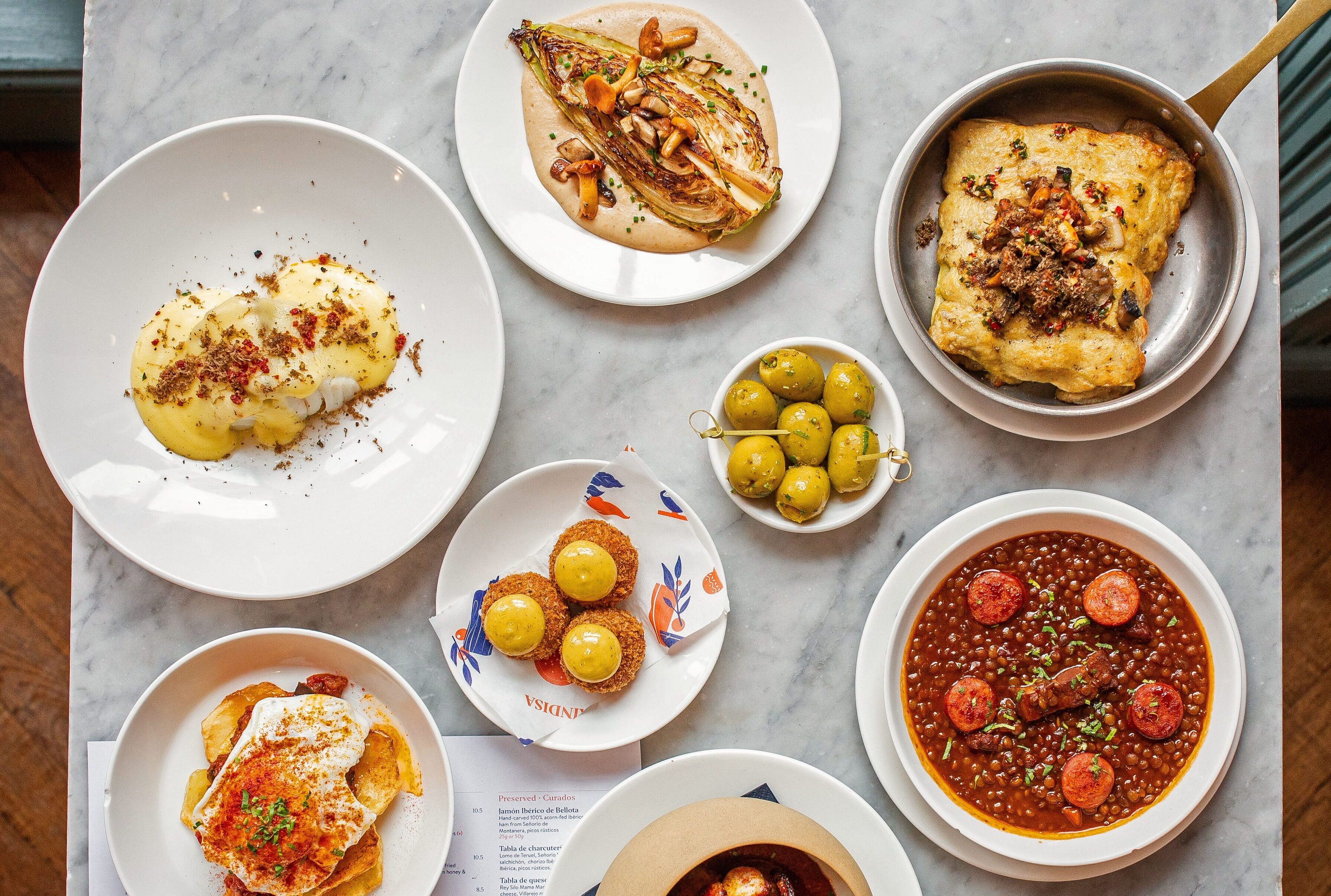 A table spread of Brindisa Tapas new Autumn dishes, including Repollo con crema de ajo blanco, Canelones de setas, Bacalao con puré de patata al azafrán, Huevos rotos with Jamón serrano, Lentejas con chorizo and Croquetas de setas