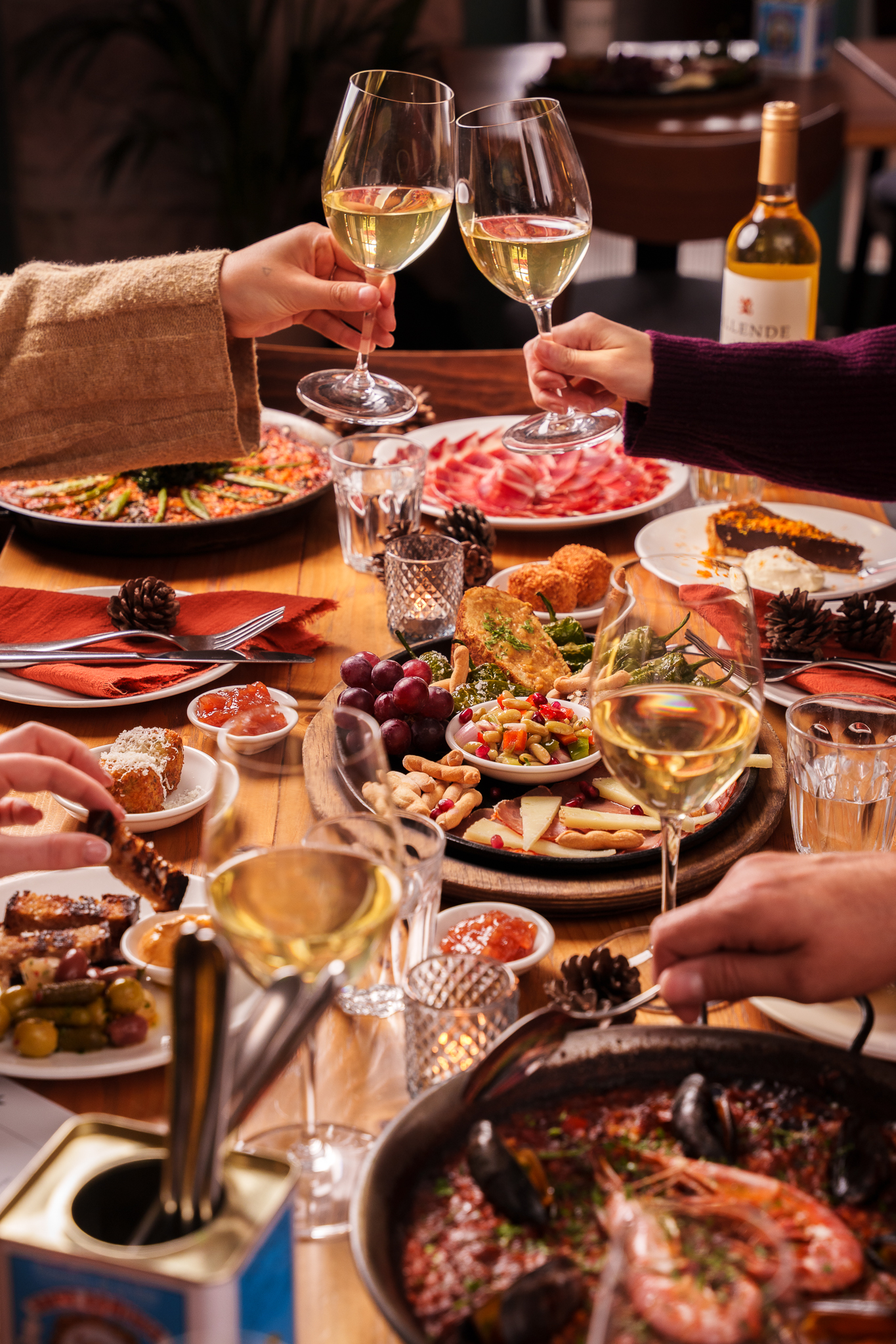 A festive table setting featuring a variety of tapas dishes, with hands raising glasses of white wine in celebration.